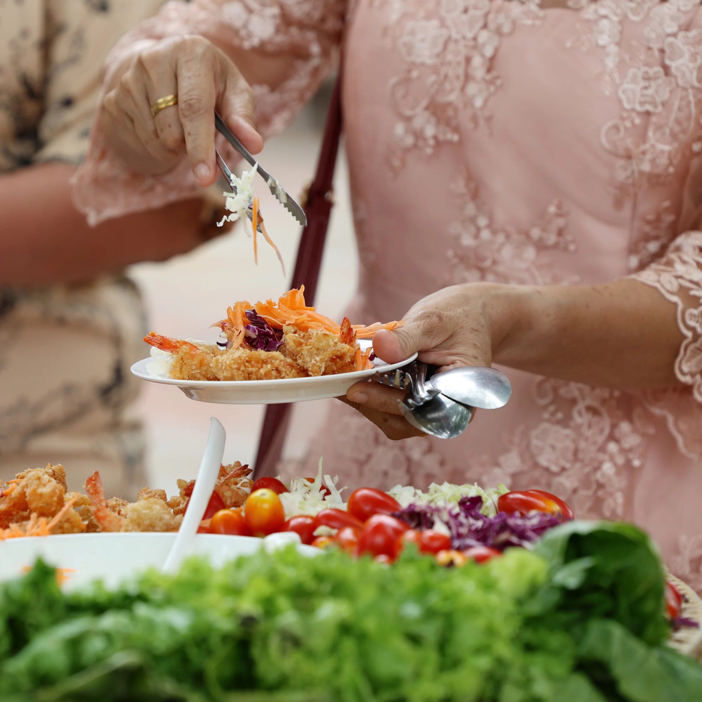 A woman in a pink lace dress serves food with tongs onto a small plate at a buffet of leafy greens, tomatoes, and shredded vegetables.