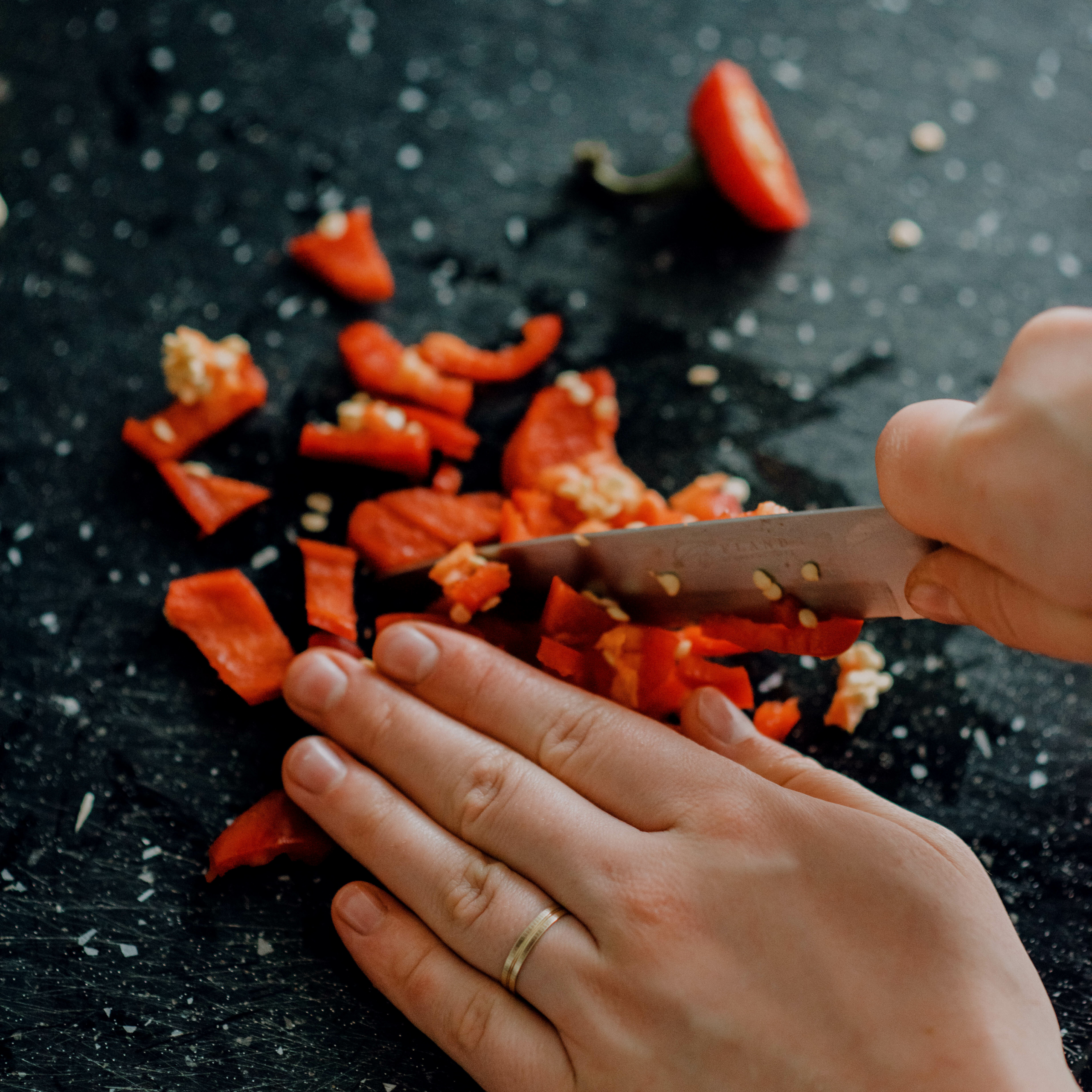 Hands chop red bell pepper on a dark, textured cutting board; scattered pieces and seeds create a vivid, focused kitchen scene.