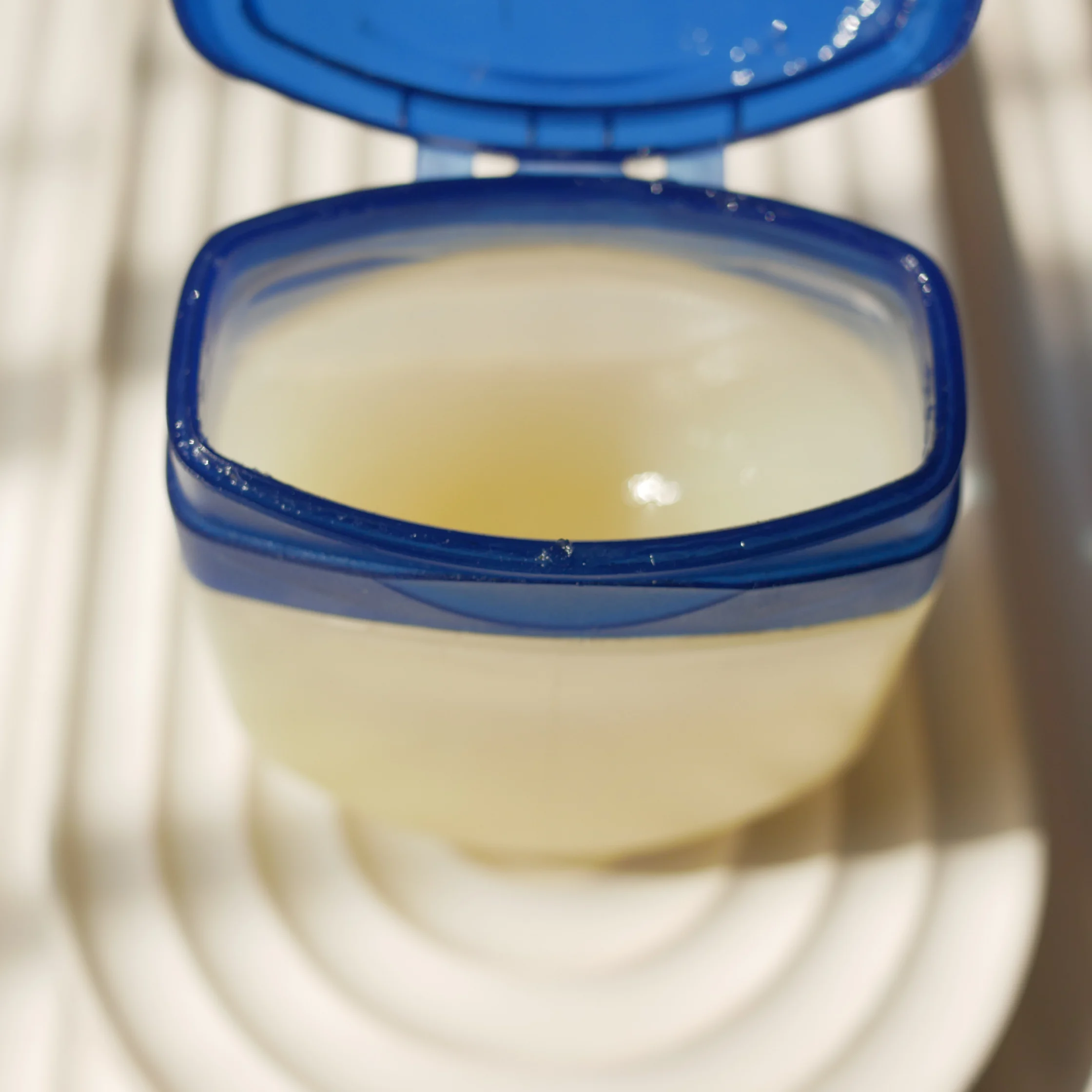 An open blue-lidded plastic container holds pale yellow liquid, resting on a white ridged surface; the close-up feels clean and clinical.