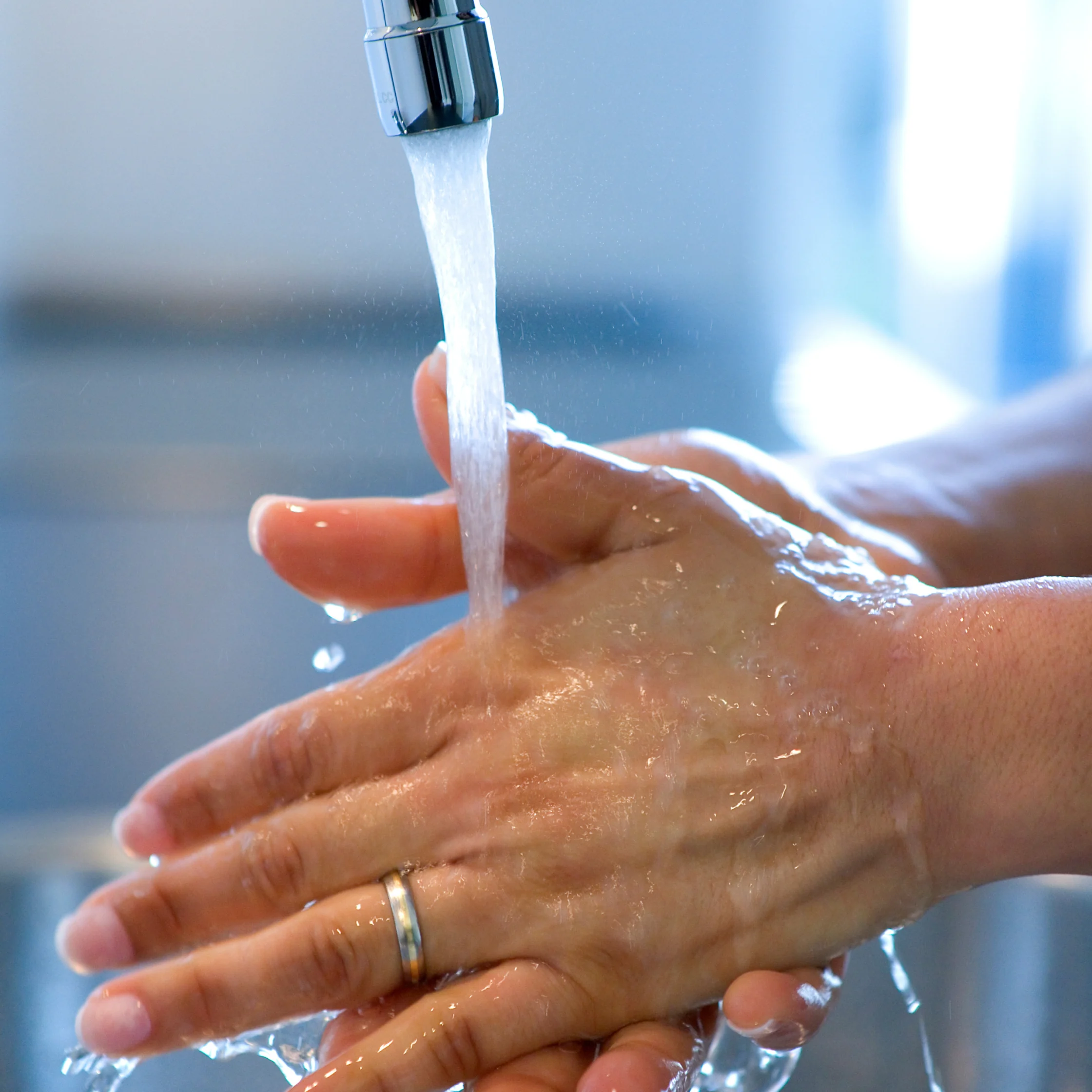 Close-up of hands with a wedding ring being washed under a running chrome faucet, water splashing over wet skin in a clean, calm kitchen setting.