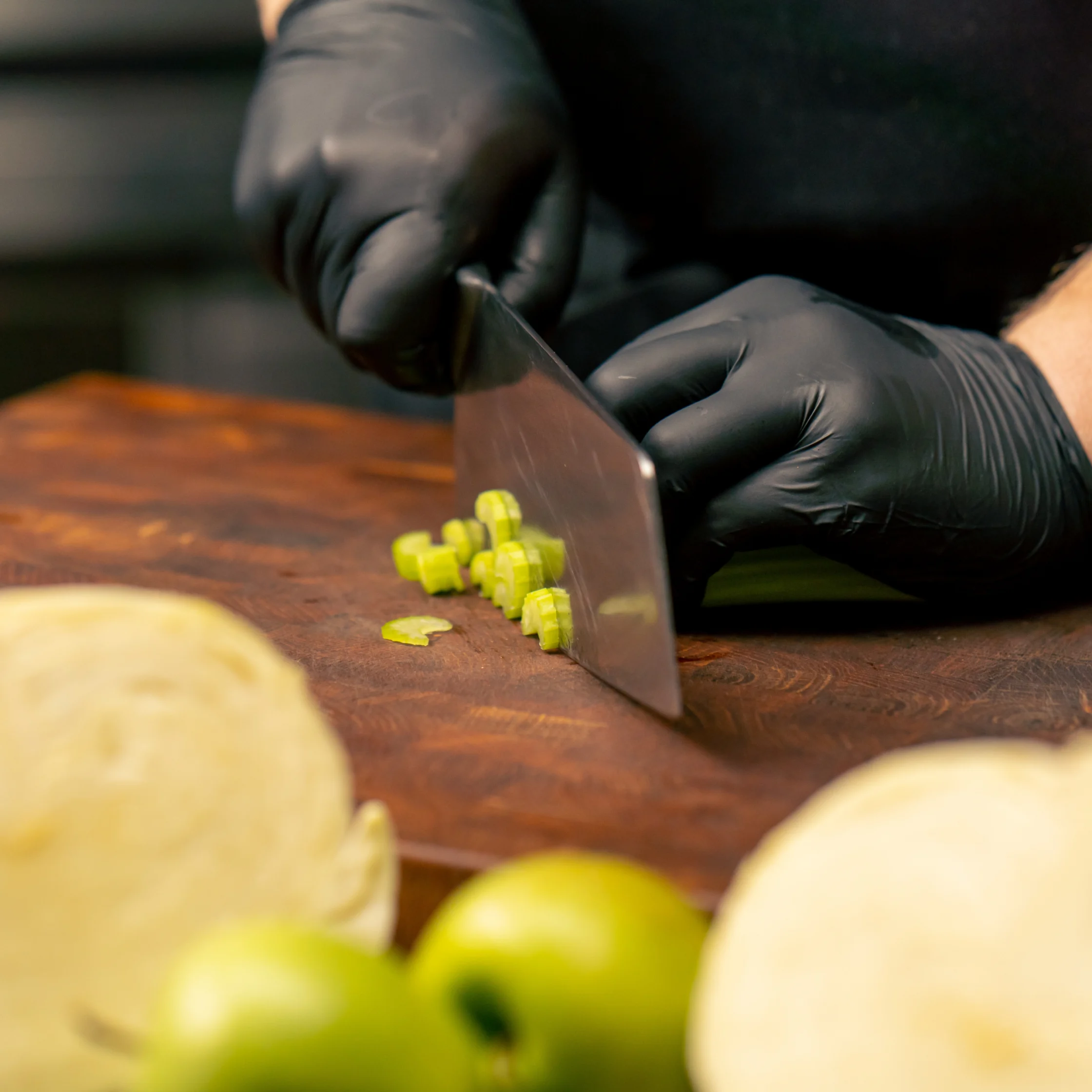 Black-gloved hands slice green onions with a chef’s knife on a wooden cutting board, with cabbage and limes nearby; focused, professional kitchen scene.