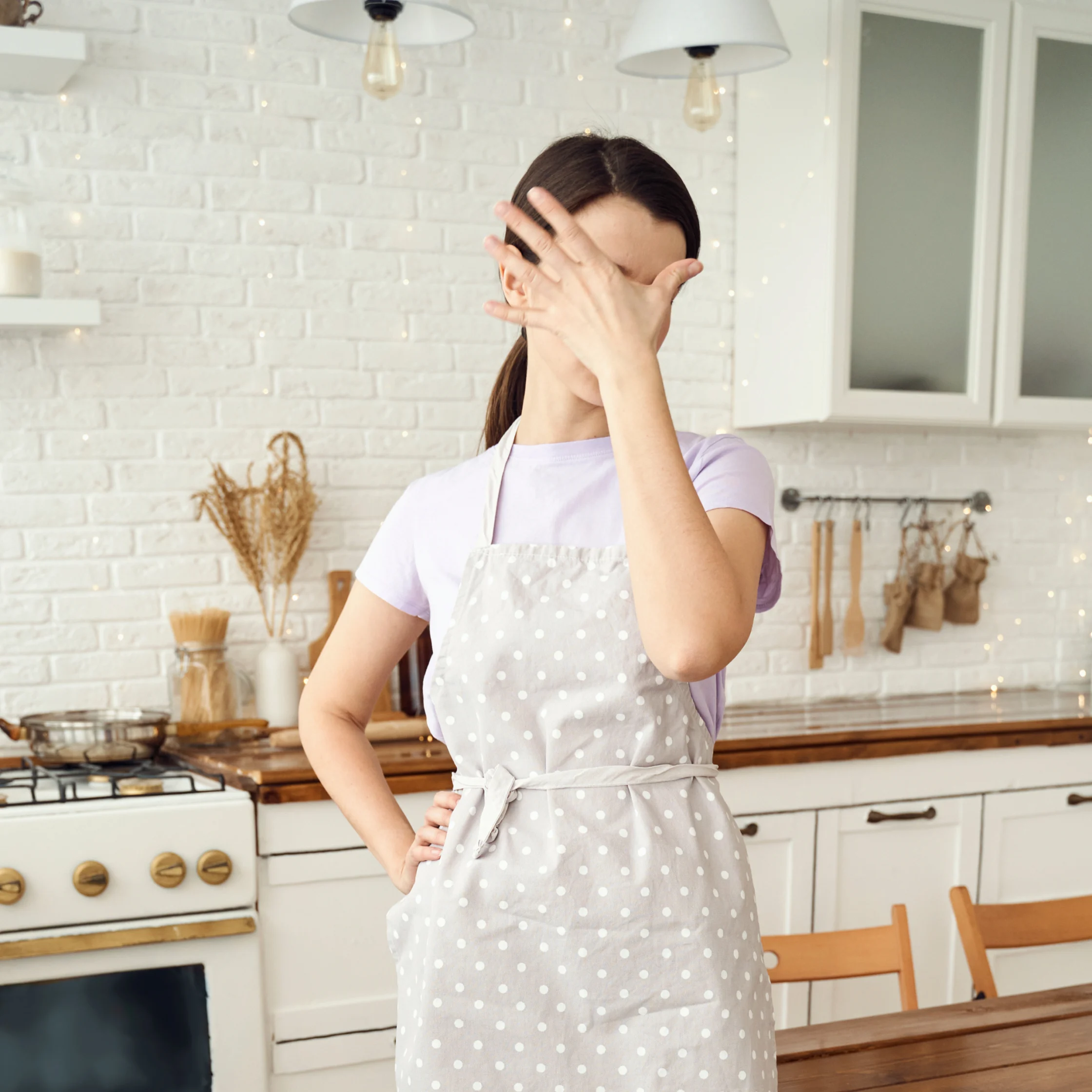 A woman in a polka-dot apron playfully covers her face in a bright, white home kitchen, creating a lighthearted, candid mood.