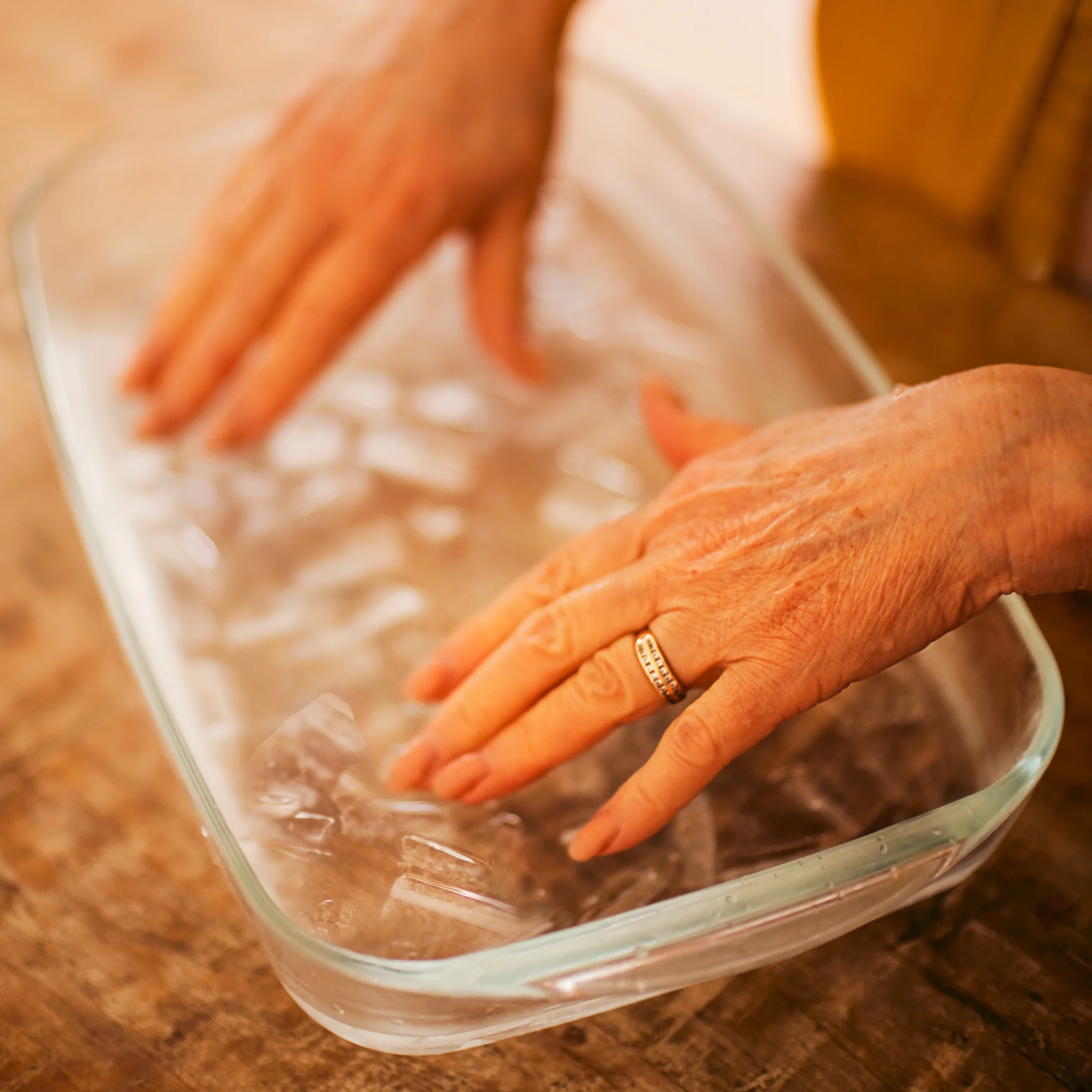 A person gently presses both hands into a clear glass dish of water on a wooden table; a gold ring catches the warm light, creating a calm, soothing mood.