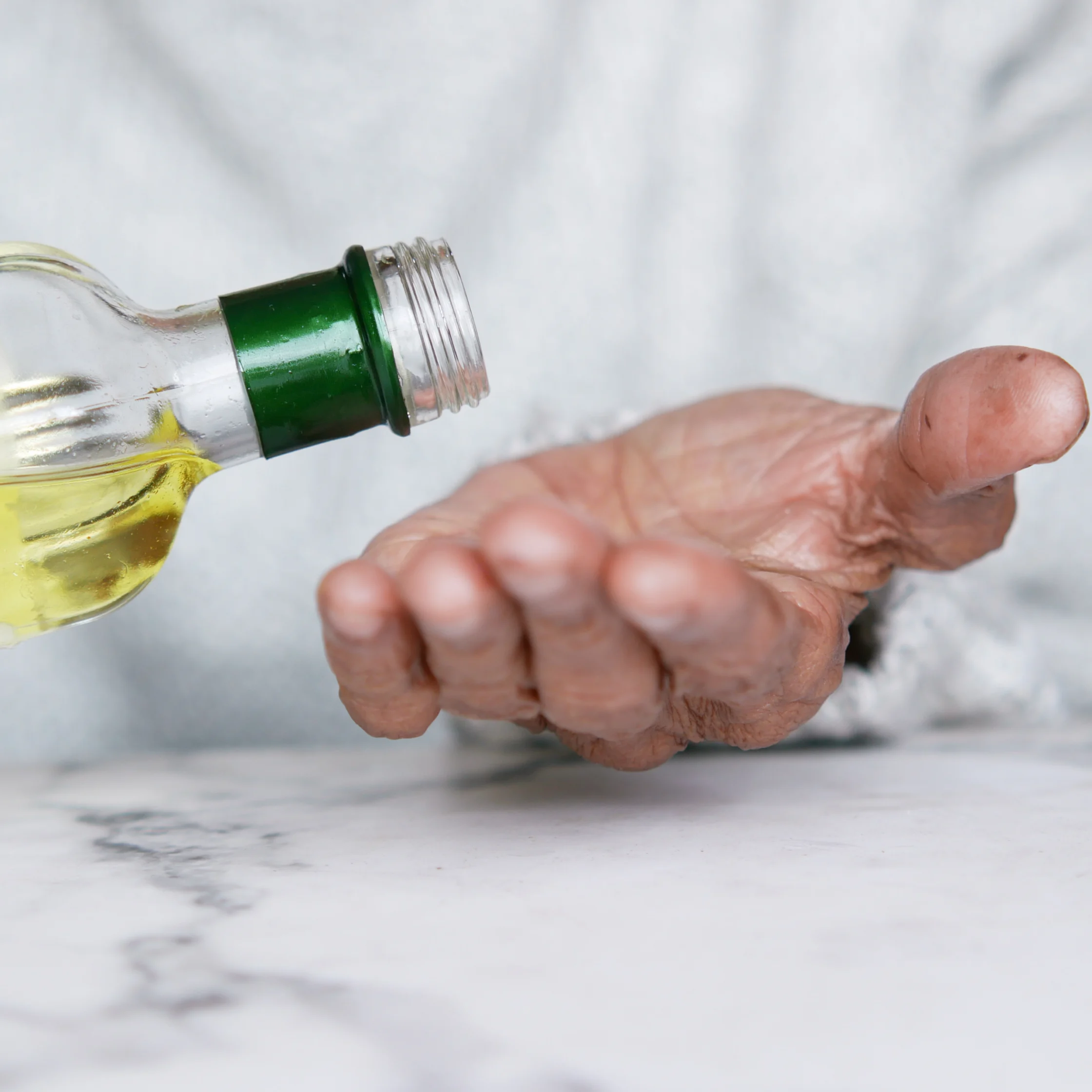 An open palm reaches beneath a glass bottle of golden oil, suggesting an offer or request; the close-up feels simple and inviting against a pale marble background.