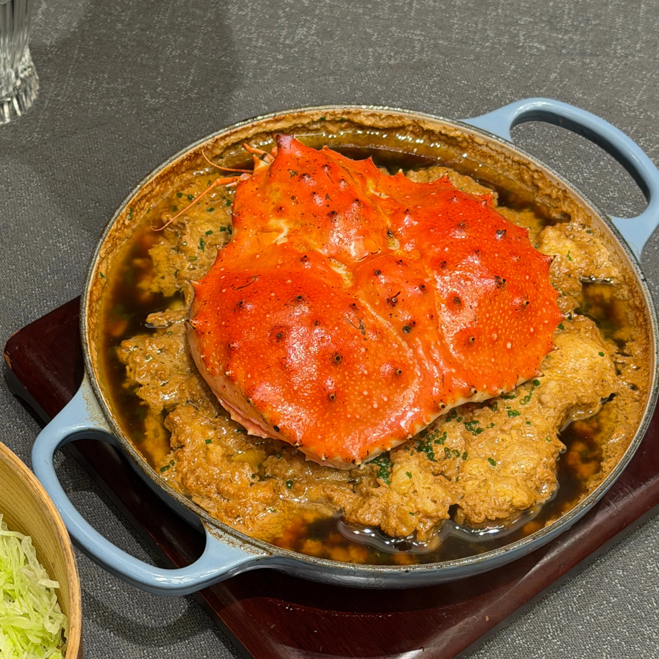 A bright red crab rests on creamy, herb-speckled seafood stew in a blue-handled pan on a wooden board, creating a rich, inviting meal.